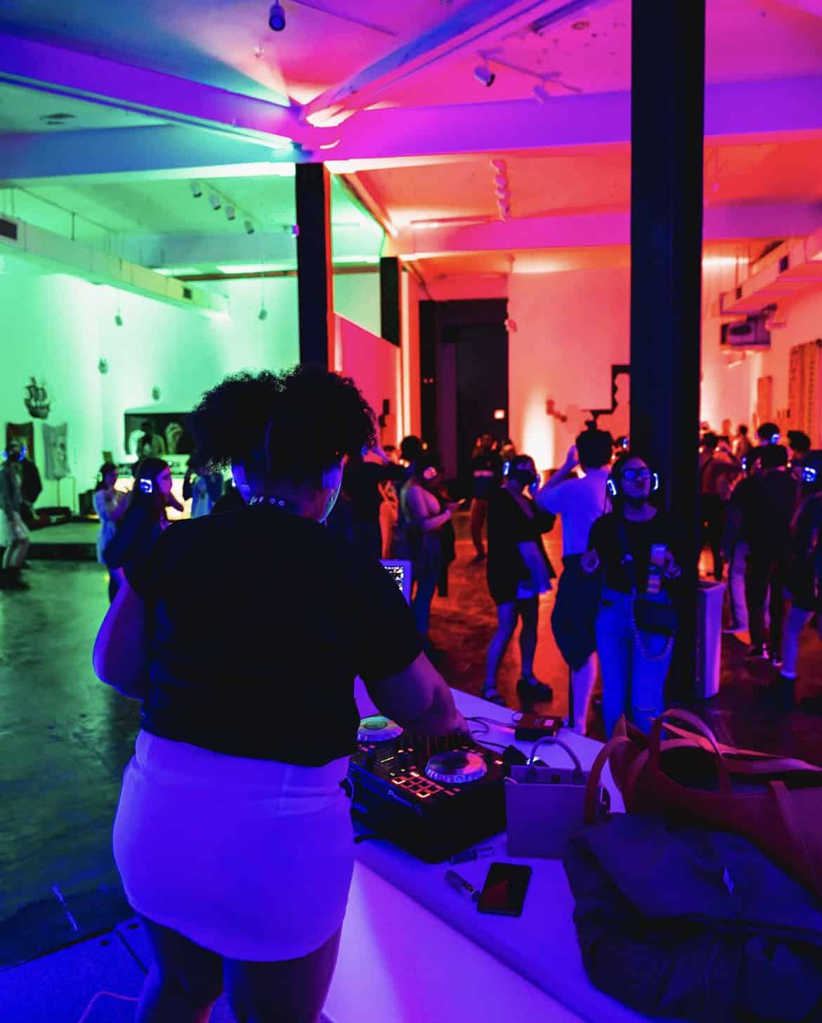A colorful photo of a silent disco at the Pittsburgh Cultural Trust. There is a DJ and people around the room wearing lit-up headphones.