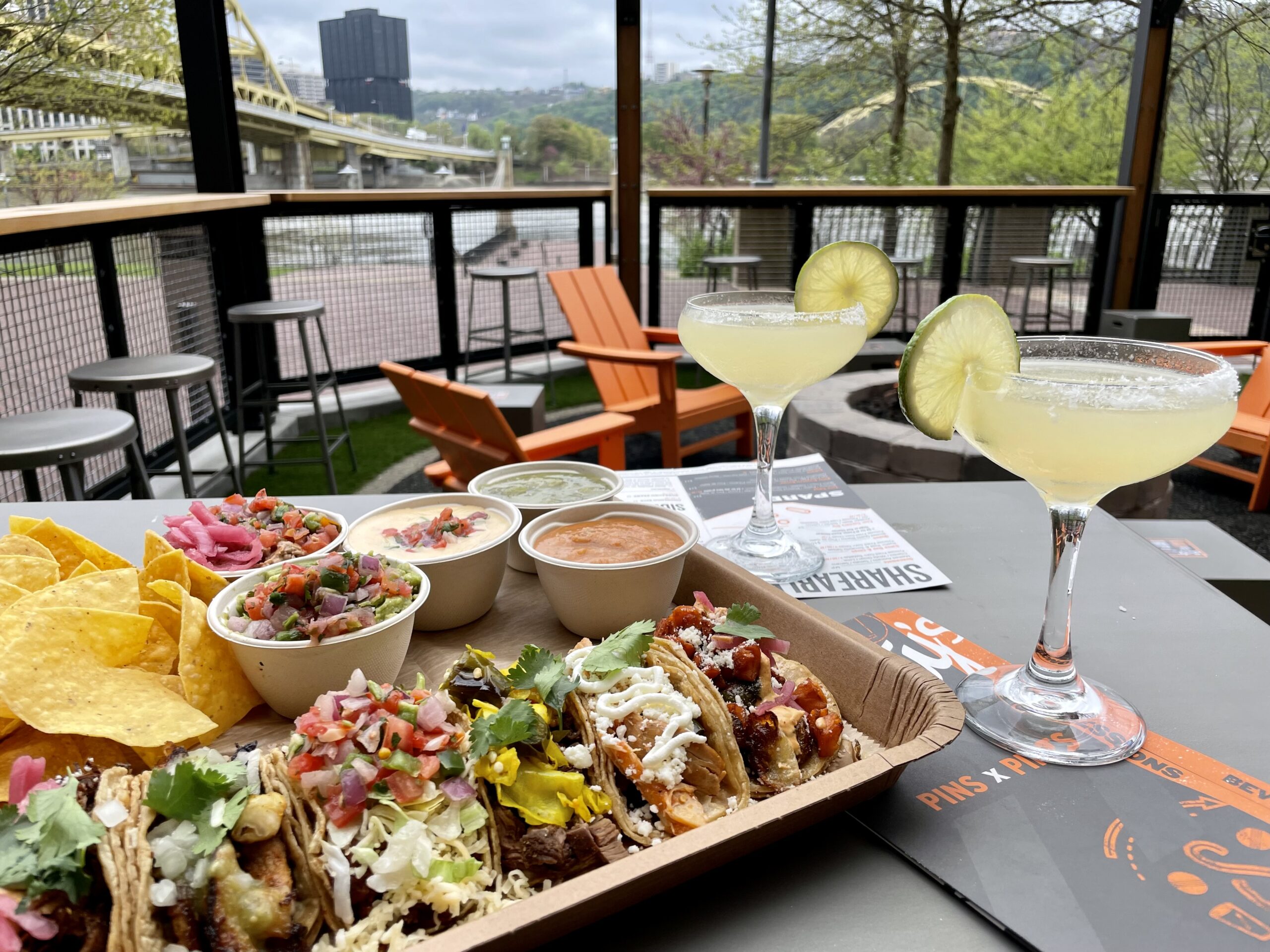 A photo at the North Shore Shorty's location, looking out at the river and bridges. There is a large assortment of tacos, dips, and drinks on the table.