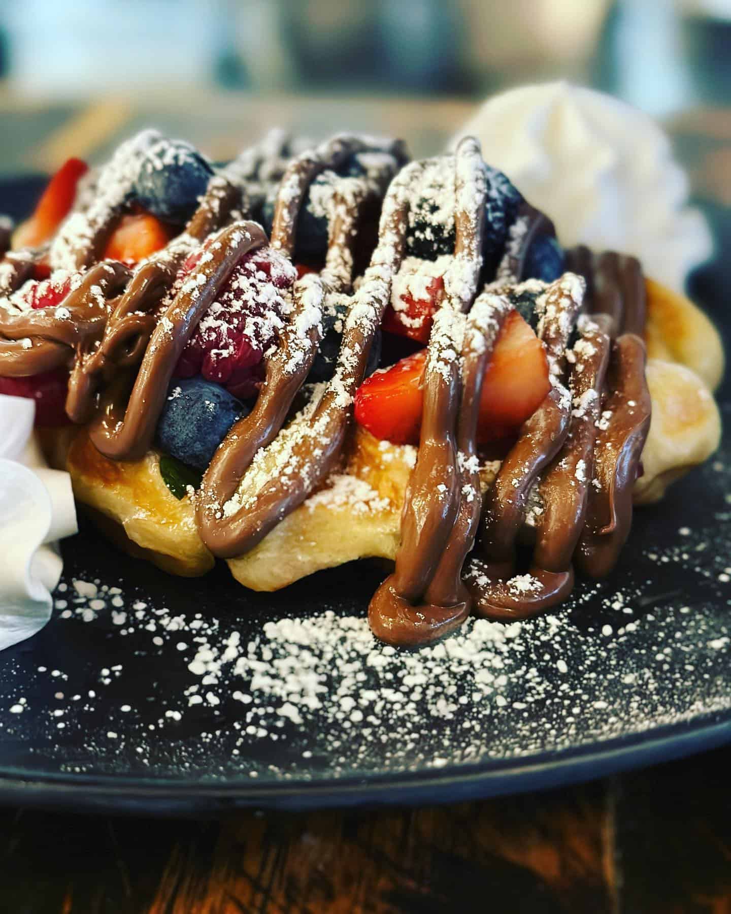 A closeup photo of a waffle with powdered sugar, fruit, and a chocolate drizzle.
