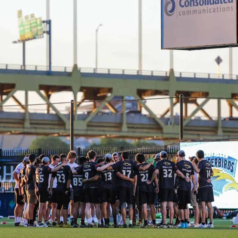 A photo of the Pittsburgh Thunderbirds team huddling at Highmark Stadium. A Pittsburgh bridge is in the background.