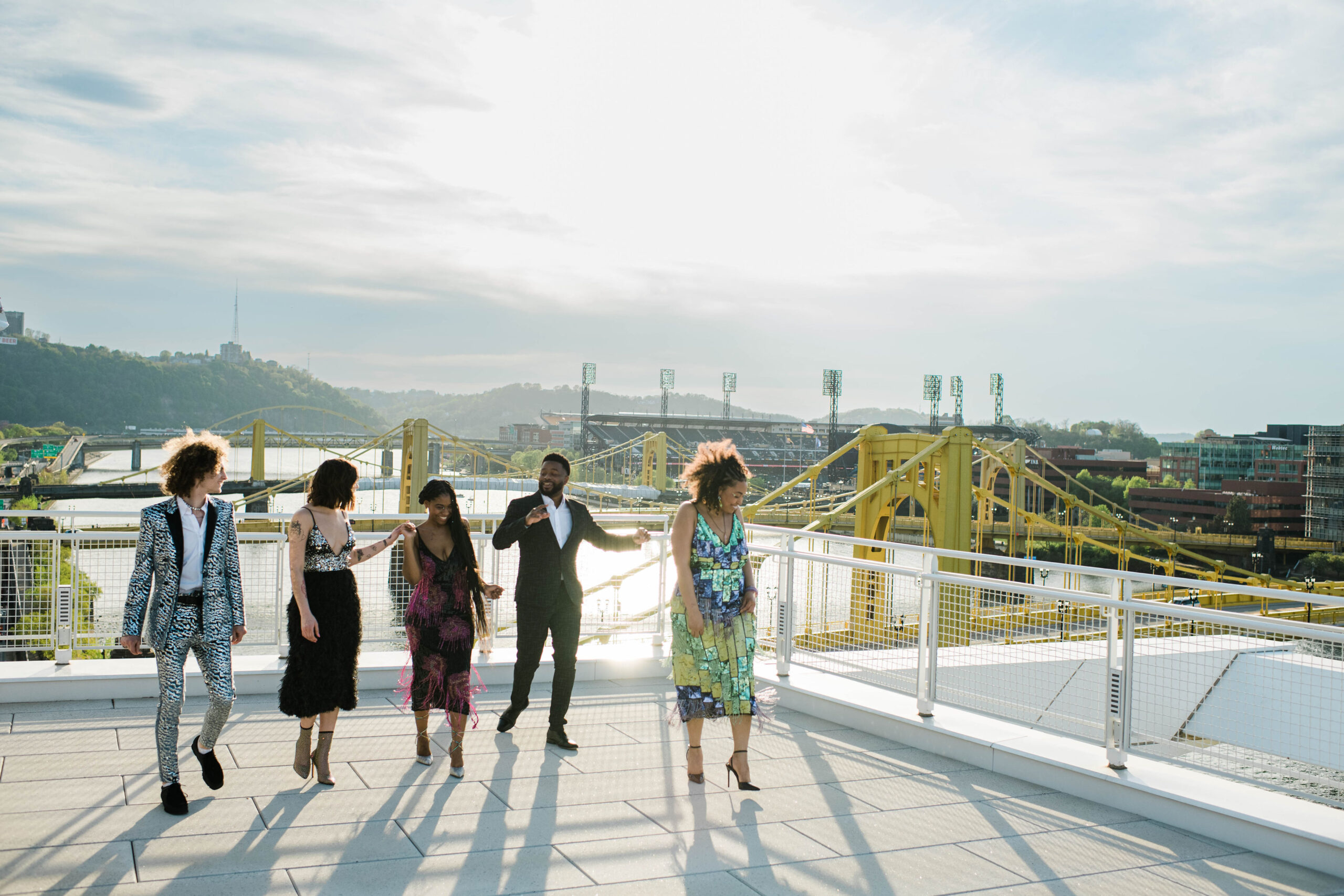 A photo of 5 people talking and dancing on the Rooftop Terrace. In the background are the 3 Sisters Bridges and PNC Park.