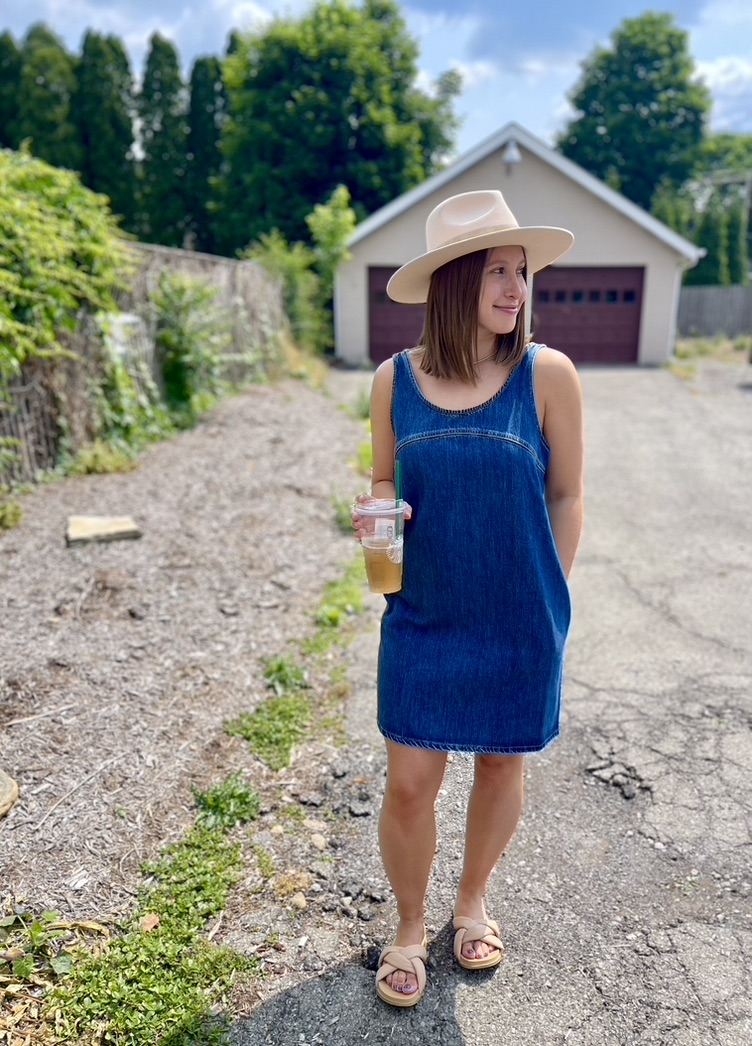 A photo of a woman in a denim dress and tan sandals with a tan hat on. She is holding a tea.