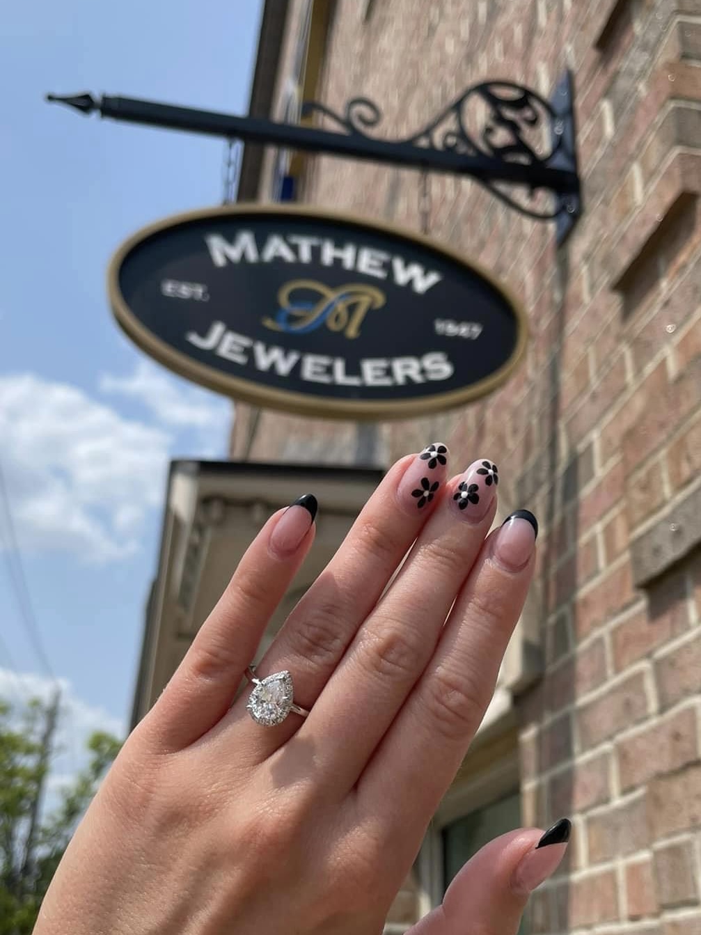 A photo of a hand with painted nails and a sparkling ring on the ring finger, with the Mathew Jewelers outdoor sign in the background.