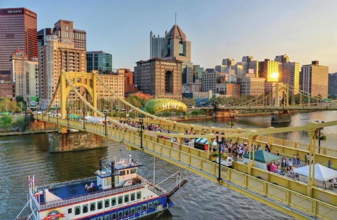 A photo of Picklesburgh on the bridge in 2022. Downtown Pittsburgh is behind the bridge and the Gateway Clipper boat is below.