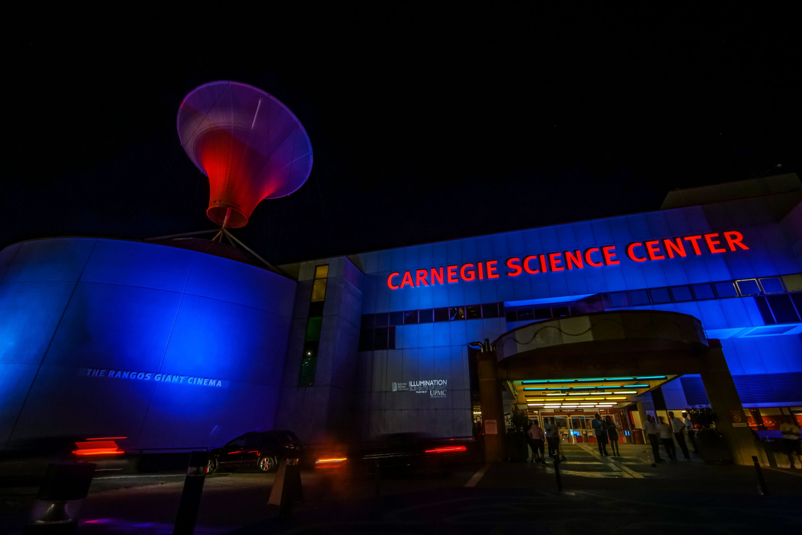 A colorful nighttime photo of the Carnegie Science Center in the evening during an event. The building is lit up with red and blue lights.