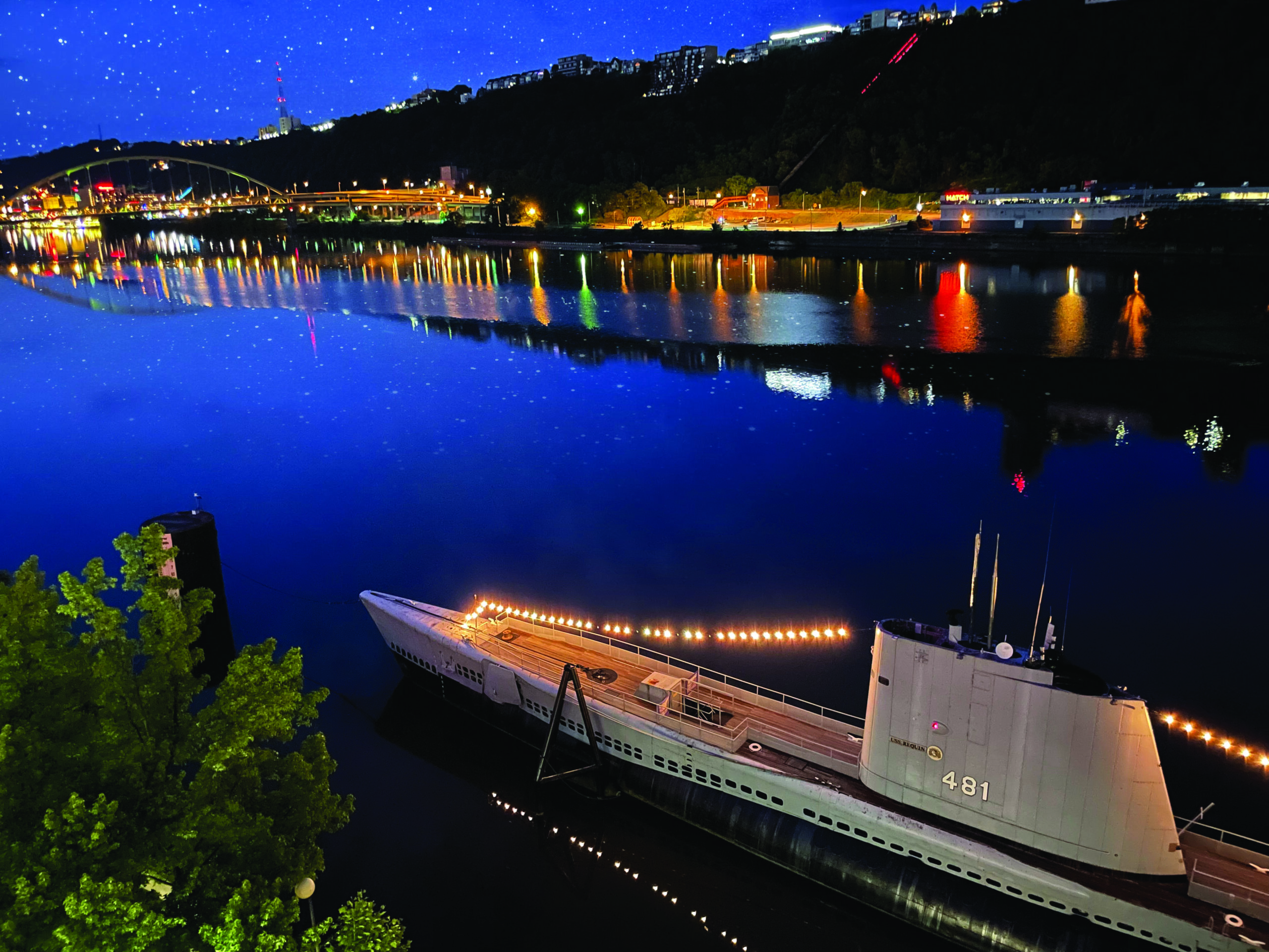 A colorful nighttime photo of the USS Requin (SS 481) Submarine on the river behind the Carnegie Science Center. Light from the bridge and buildings reflects off of the river.