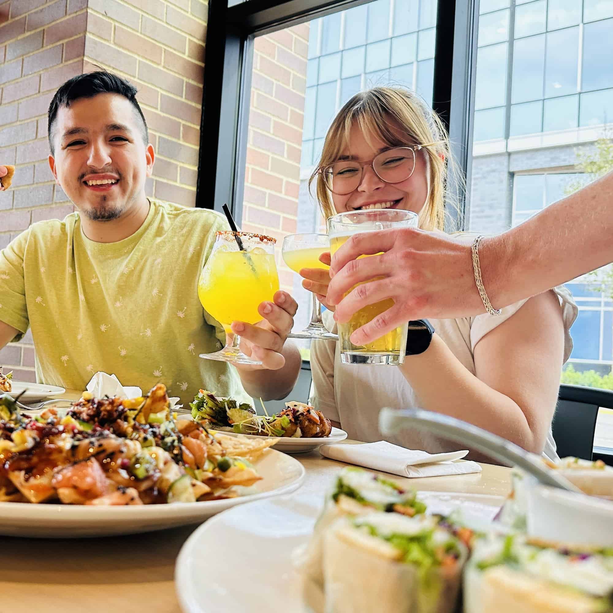 A photo of a man and a woman and a third person off camera clinking glasses together and smiling. Their table has a variety of food on it.