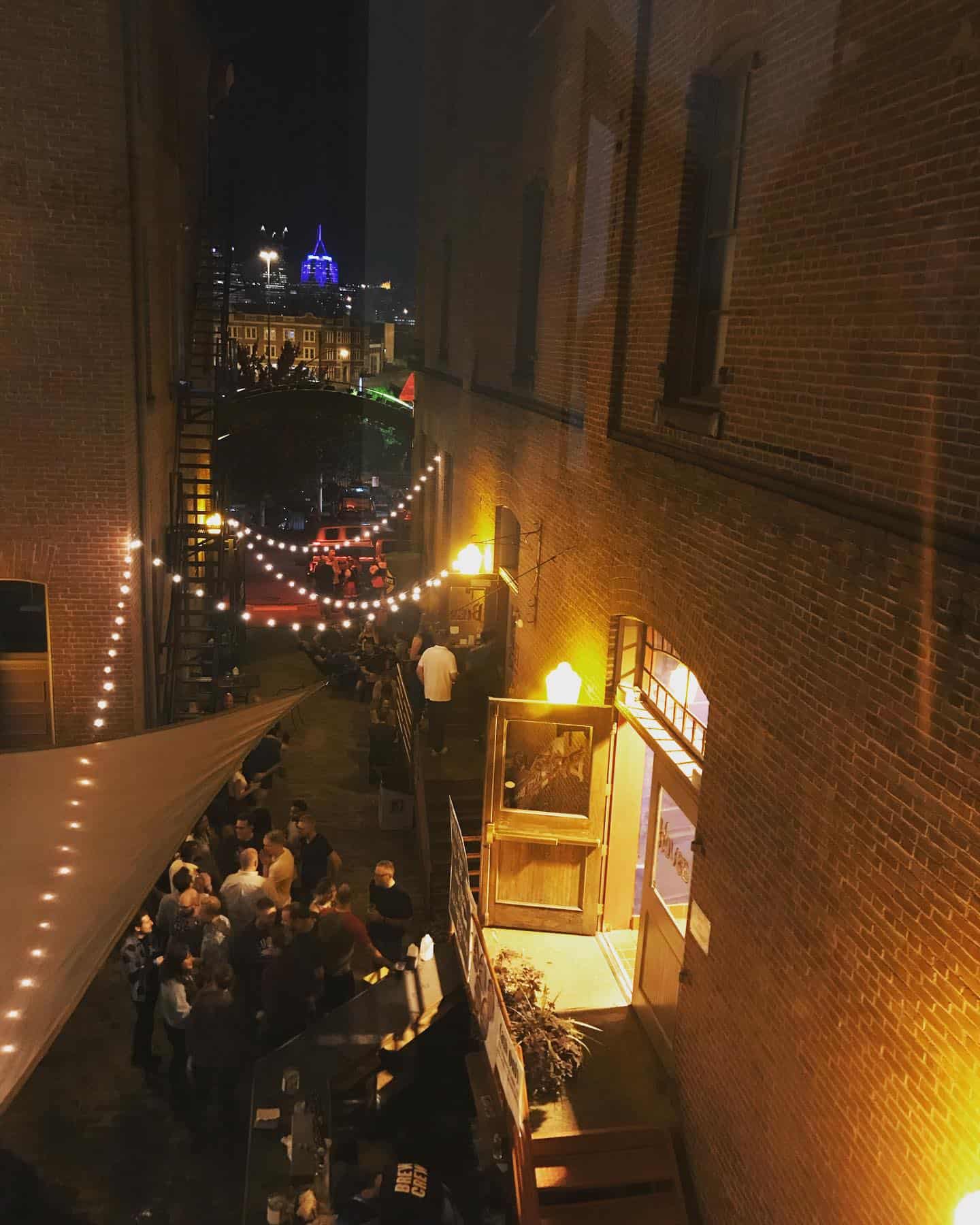 A photo of an alleyway at night with lights and people talking. Downtown Pittsburgh can be seen in the distance.