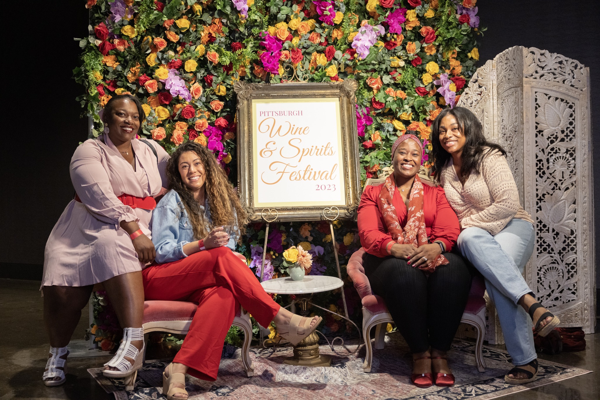 A photo of four women posing with a beautiful backdrop of flowers behind them. The sign in front of the backdrop says Wine & Spirit Festival 2023.