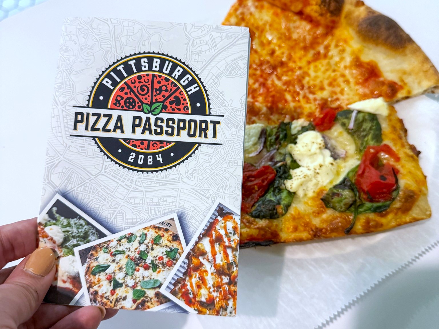 The Pittsburgh Pizza Passport book held up next to two slices of unique pizzas.