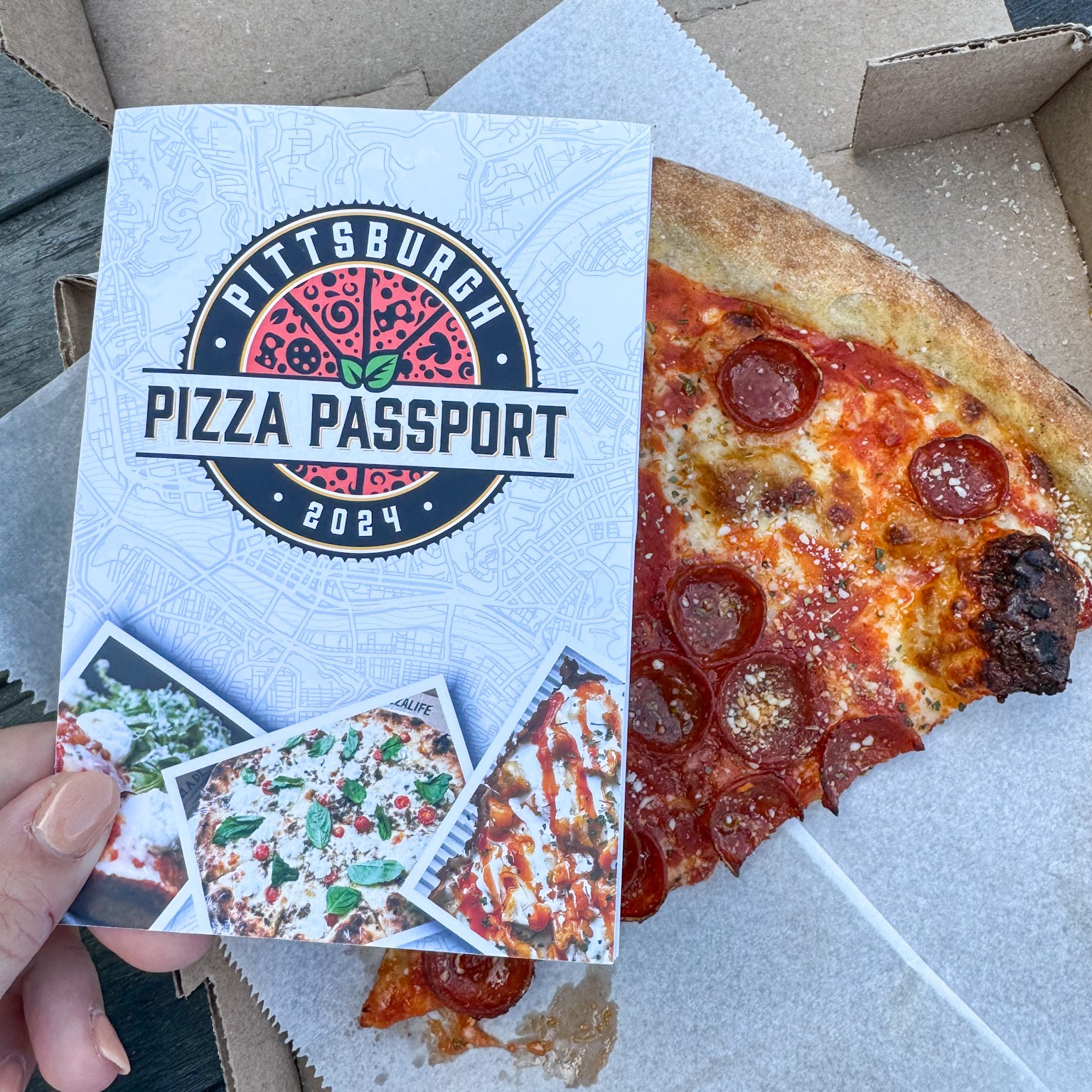 The Pittsburgh Pizza Passport book held up next to a slice of pizza from Pizzeria Davide.