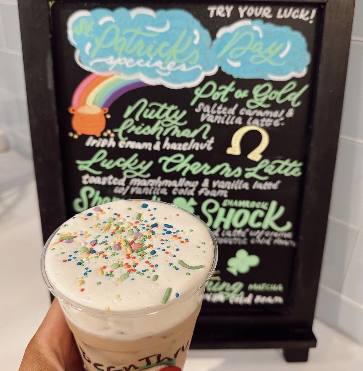 A photo of someone holding a BeanThru St. Patrick's Day themed iced coffee in front of a sign describing their holiday specials.