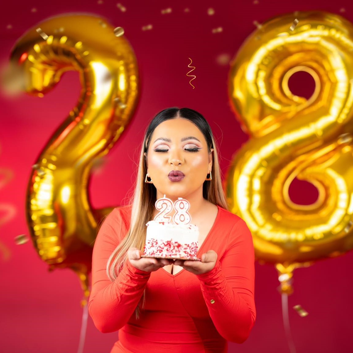 A woman blowing out "28" candles on a cake in front of "28" balloons. 