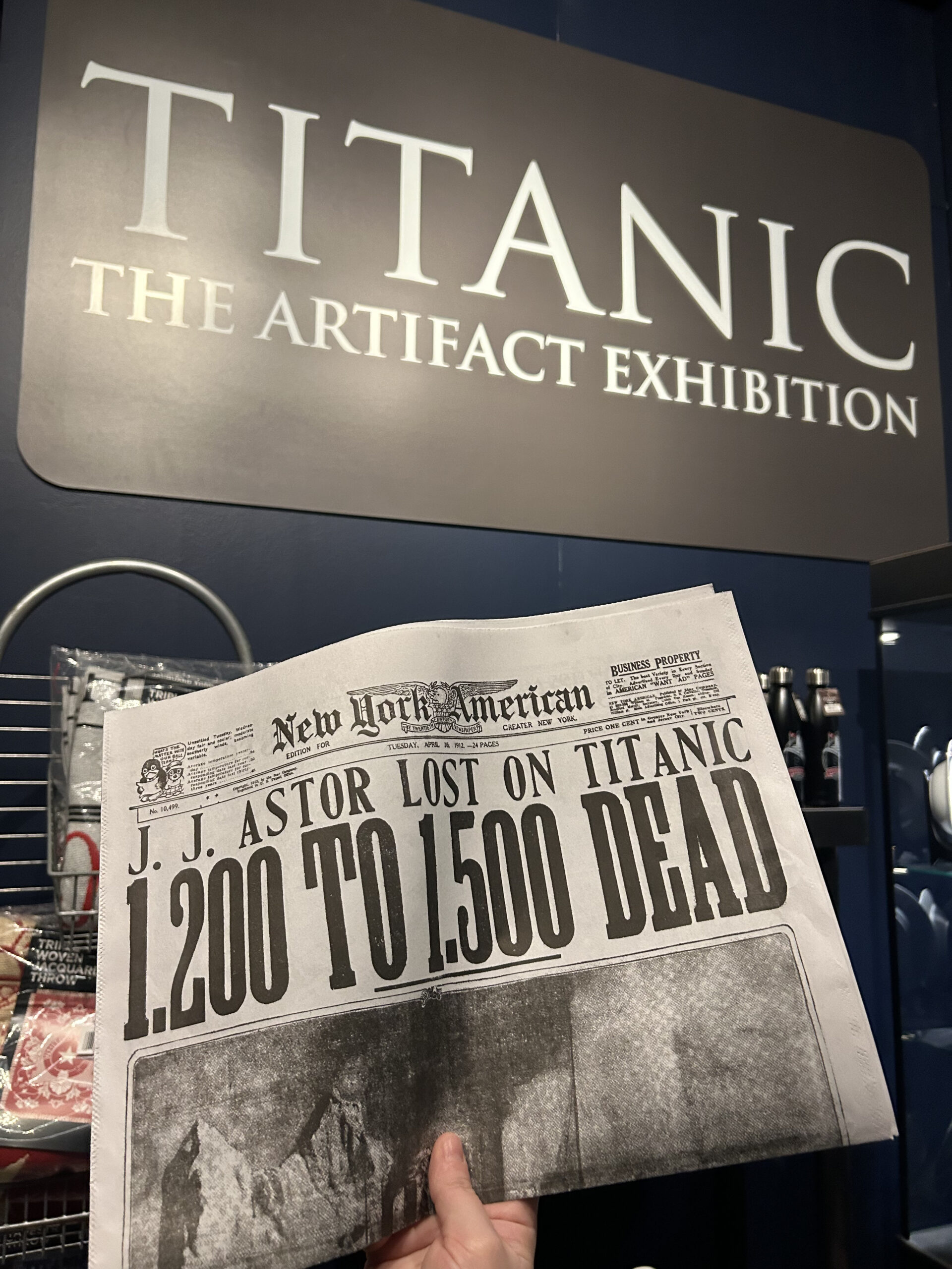 A photo of someone holding up a replica newspaper up next to the "TITANIC: THE ARTIFACT EXHIBITION" sign.