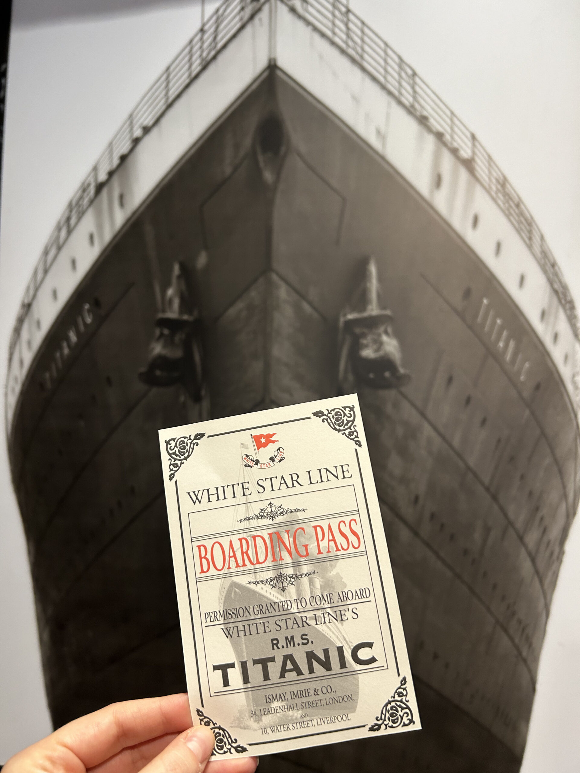 A photo of someone holding up a replica White Star Line boarding pass in front of a black and white photo of the Titanic ship.
