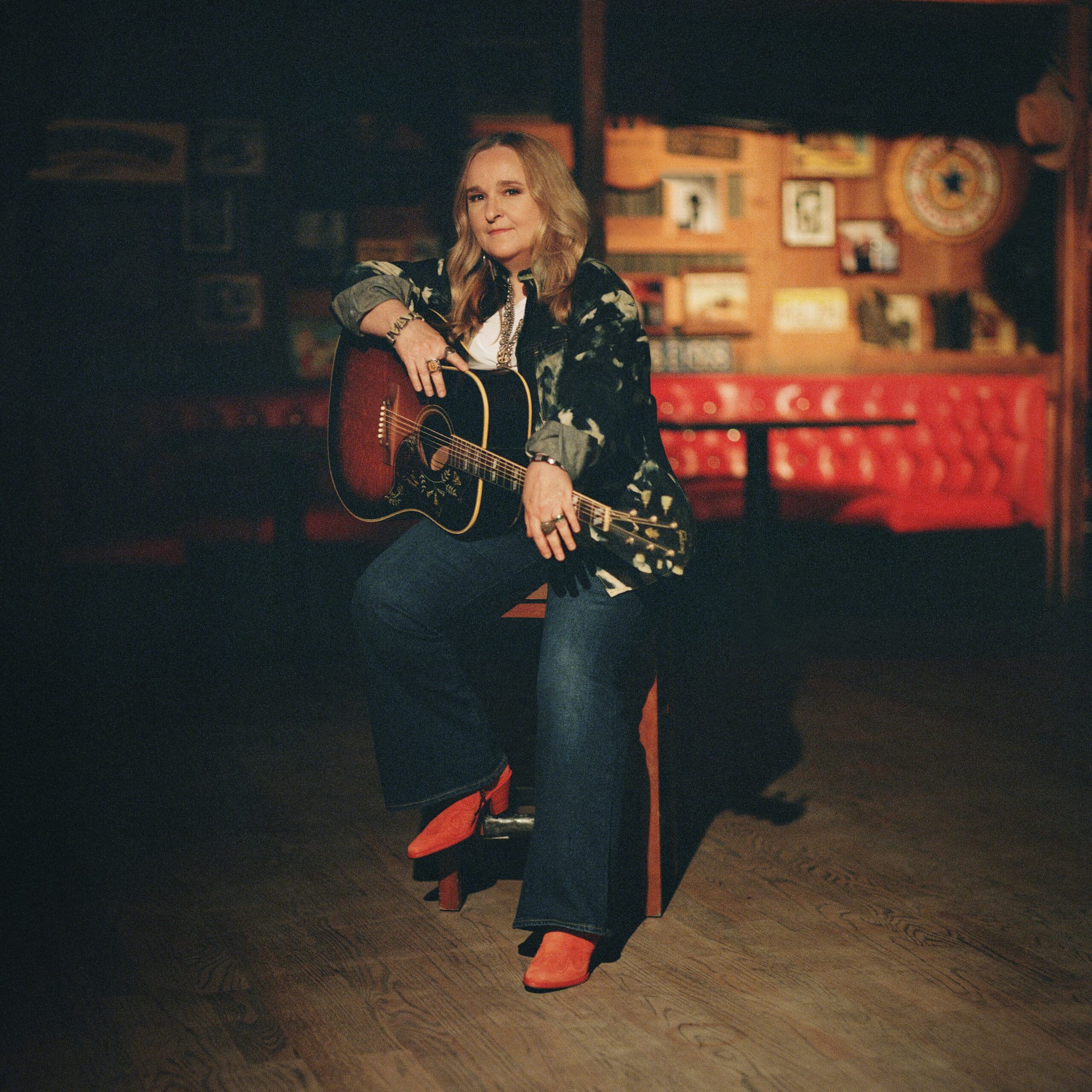 A photo of Melissa Etheridge sitting on a stool with an acoustic guitar.