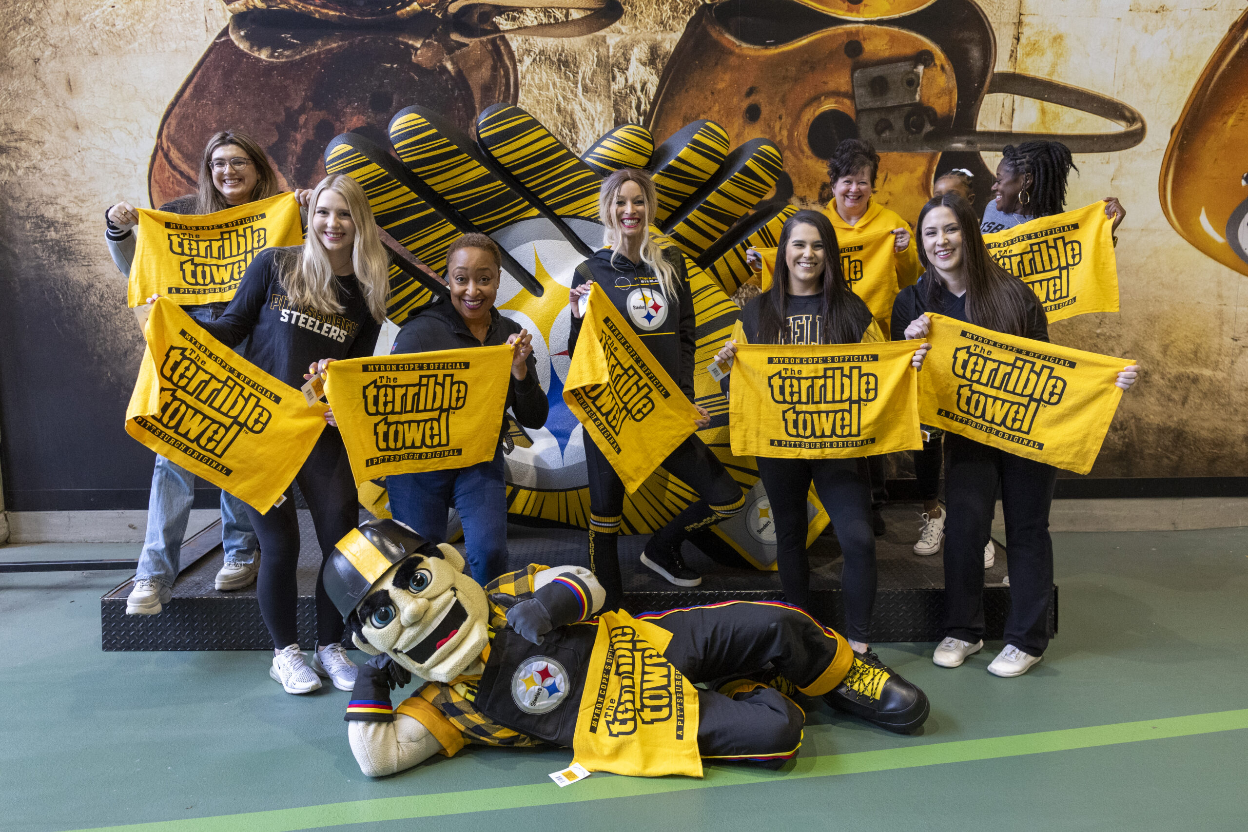 A photo of eight women and a child posing with Terrible Towels in front of the Steelers gloves at Acrisure Stadium. Steely McBeam lays on the floor posing in front of them.