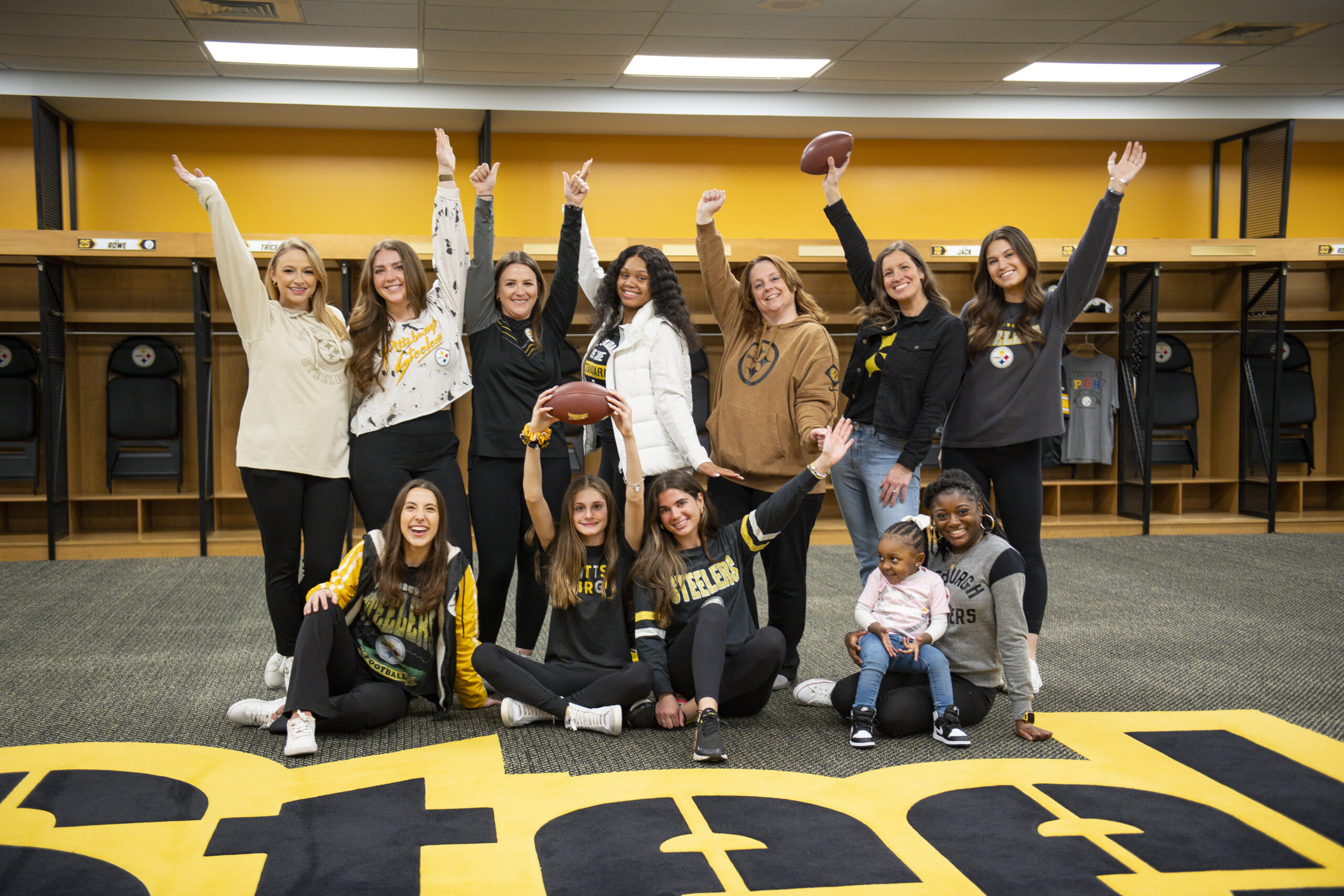 A photo of eleven women and a young girl posing with footballs and Steelers gear in the Steelers locker room at Acrisure Stadium.