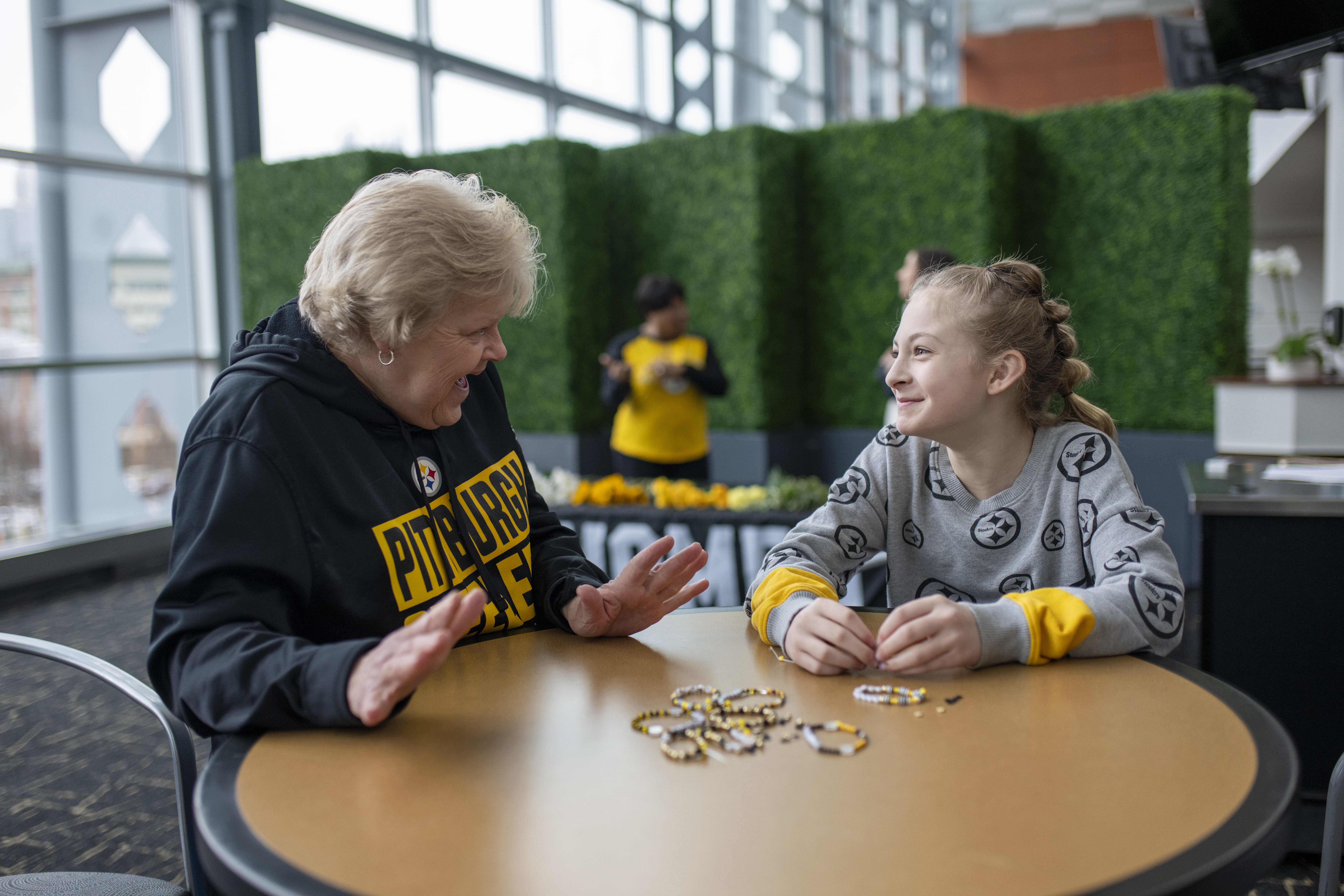 A photo of an older woman and a younger woman making black and gold friendship bracelets together at SteelHERS Social.