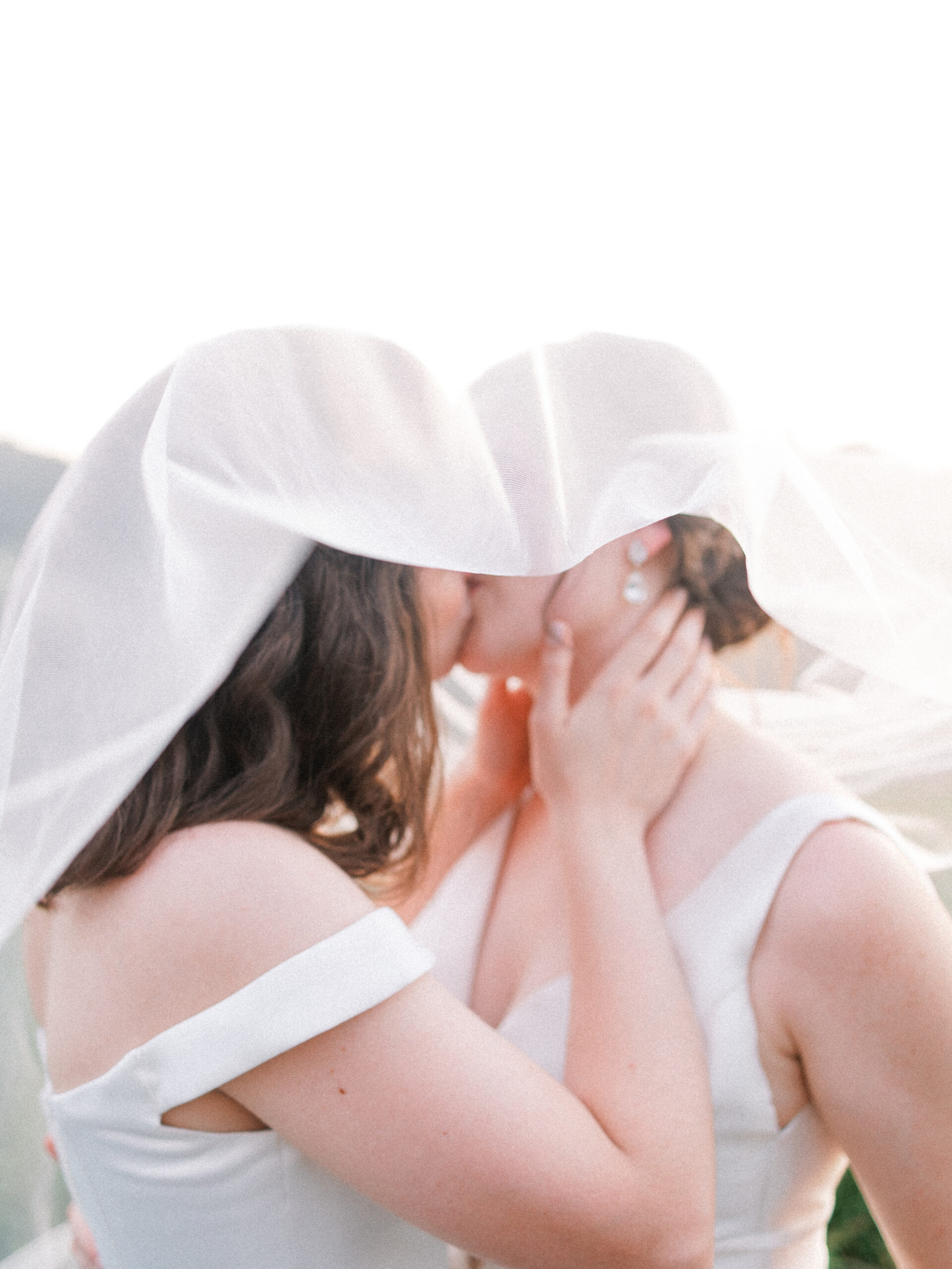 A photo of two brides kissing under a flowing veil.