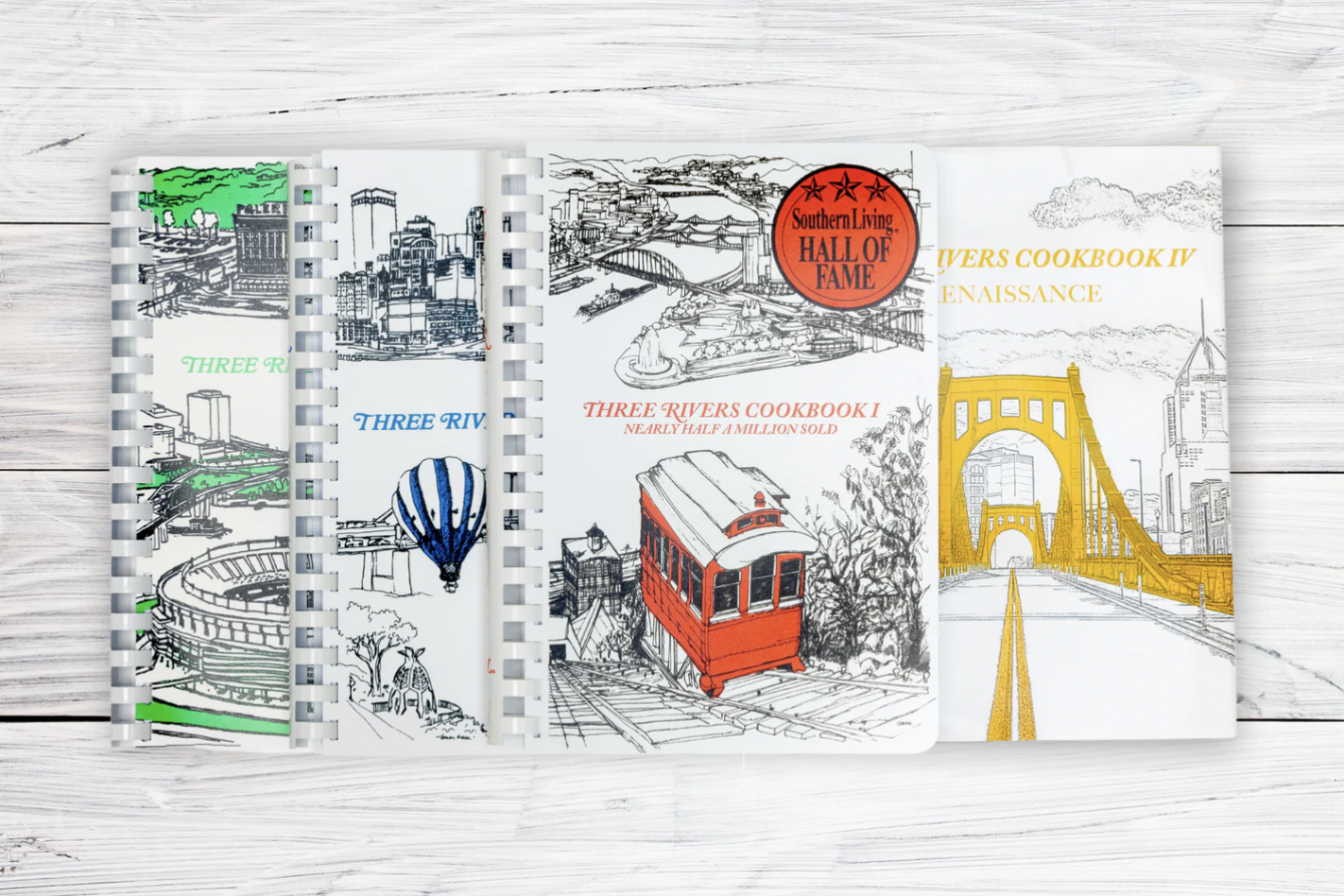 A photo of the four Three Rivers Cookbooks laid out on a white table. They are black and white covers with splashes of color in Pittsburgh illustrations, like a yellow bridge.