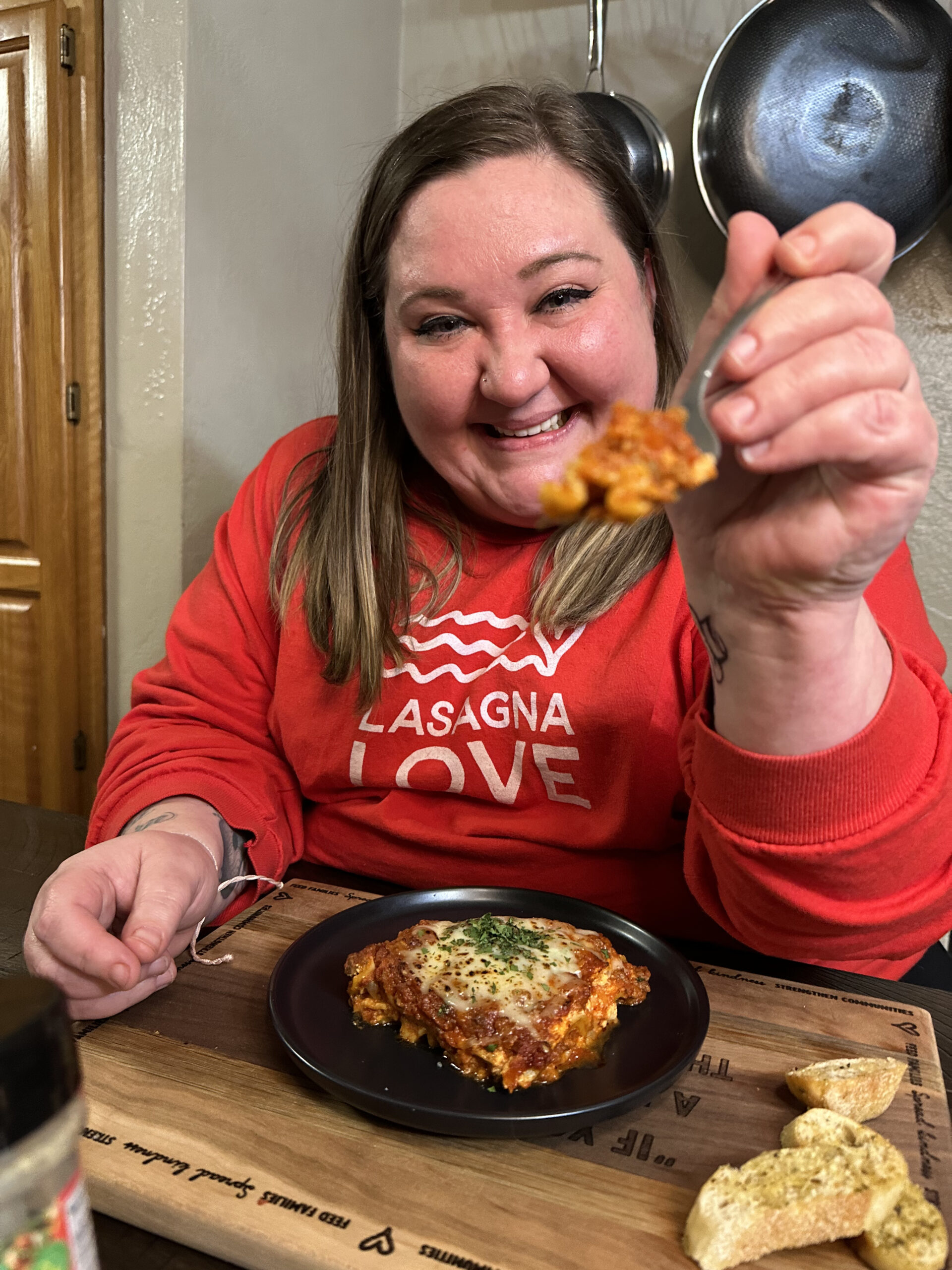 A photo of Desarae Legros holding a fork with lasagna on it up to the camera. She is wearing a red Lasagna Love shirt.
