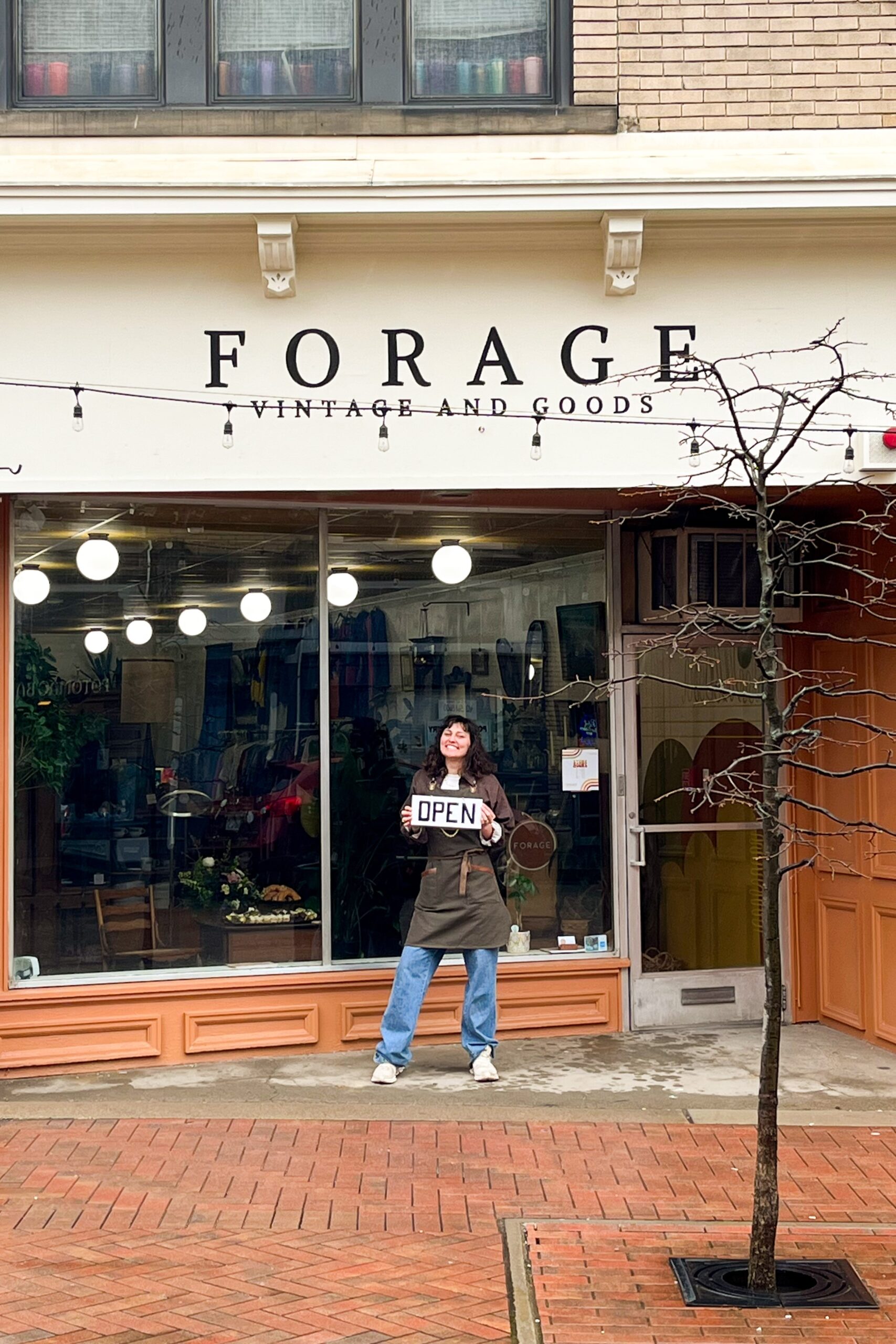 A photo of someone standing outside the Forage Vintage and Goods storefront in Dormont, holding an OPEN sign and smiling.