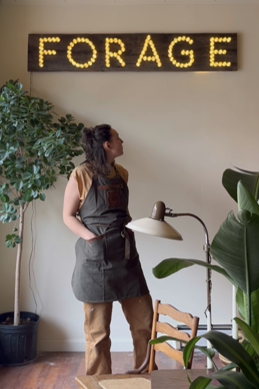A photo of someone in a working apron looking back at a wall that has a FORAGE sign with lights.