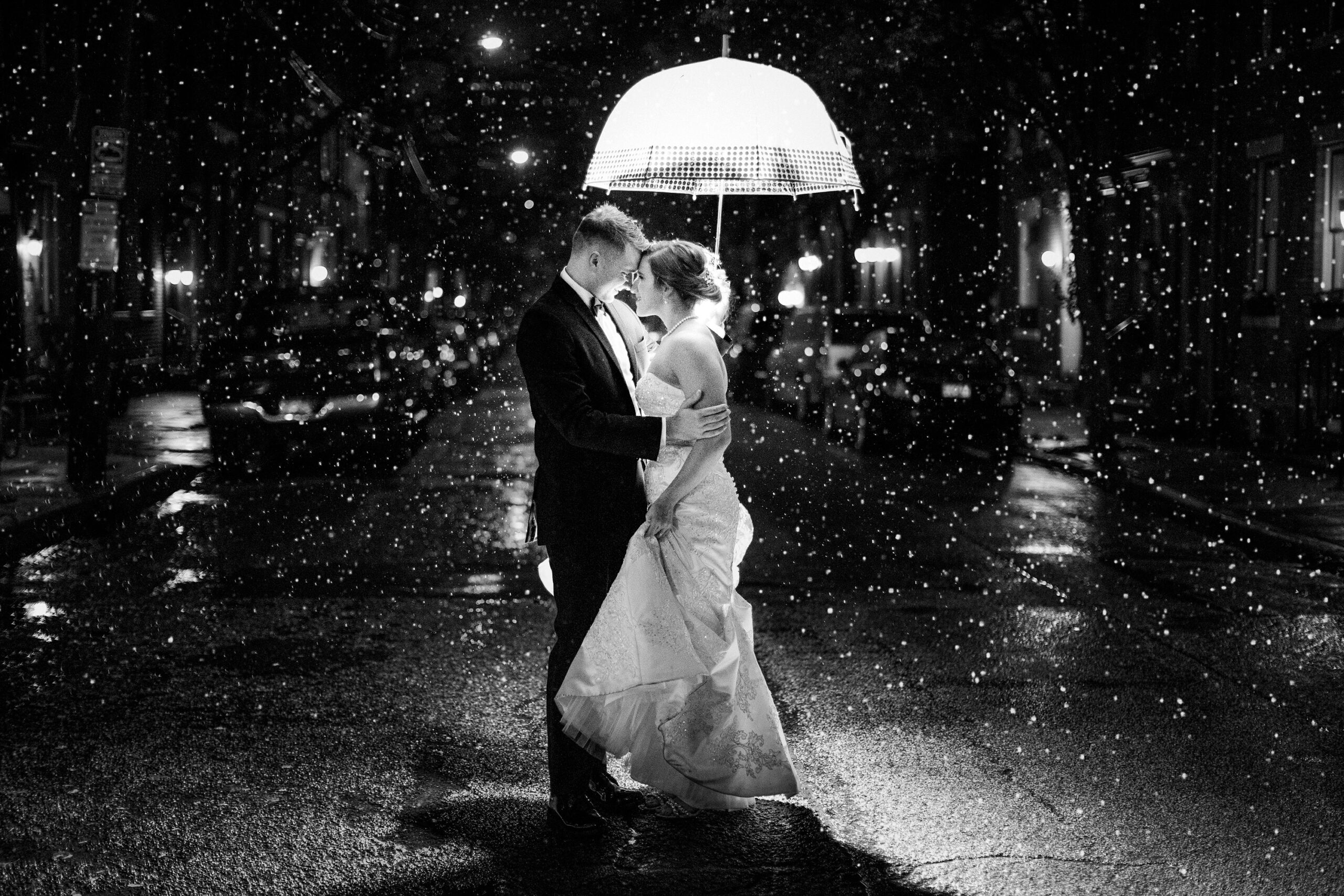 A black and white dramatic photo of a bride and groom on a street under an umbrella.