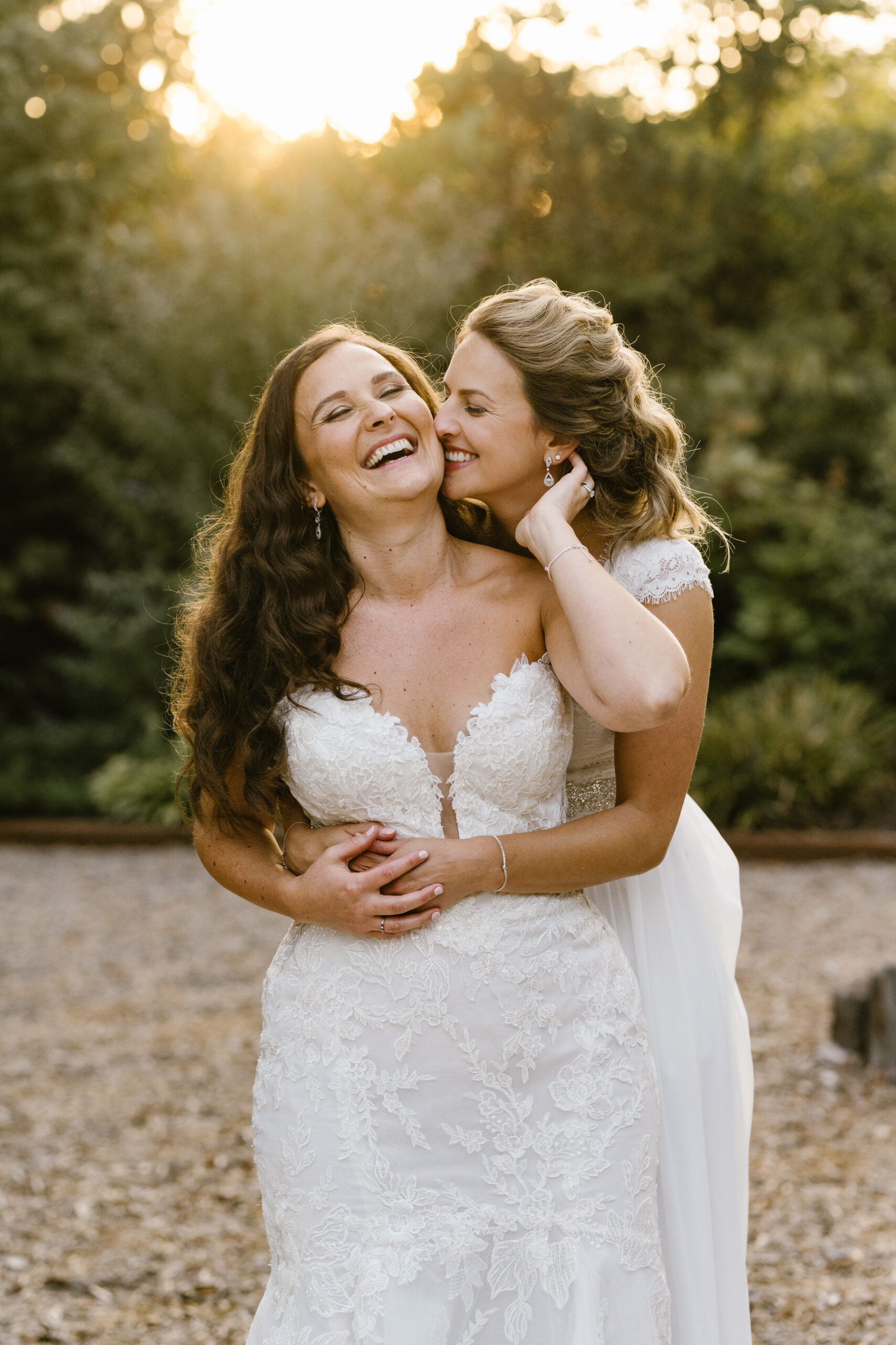 A bride holds a bride from behind while they're both smiling and laughing.