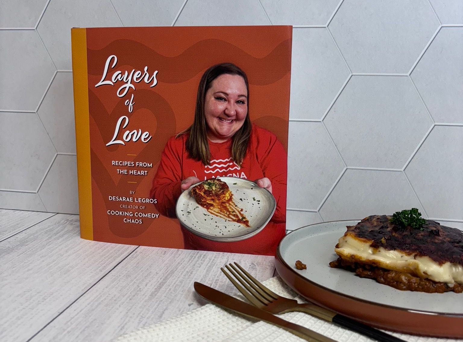 A photo of the Layers of Love book by Desarae Legros next to a plate of lasagna.