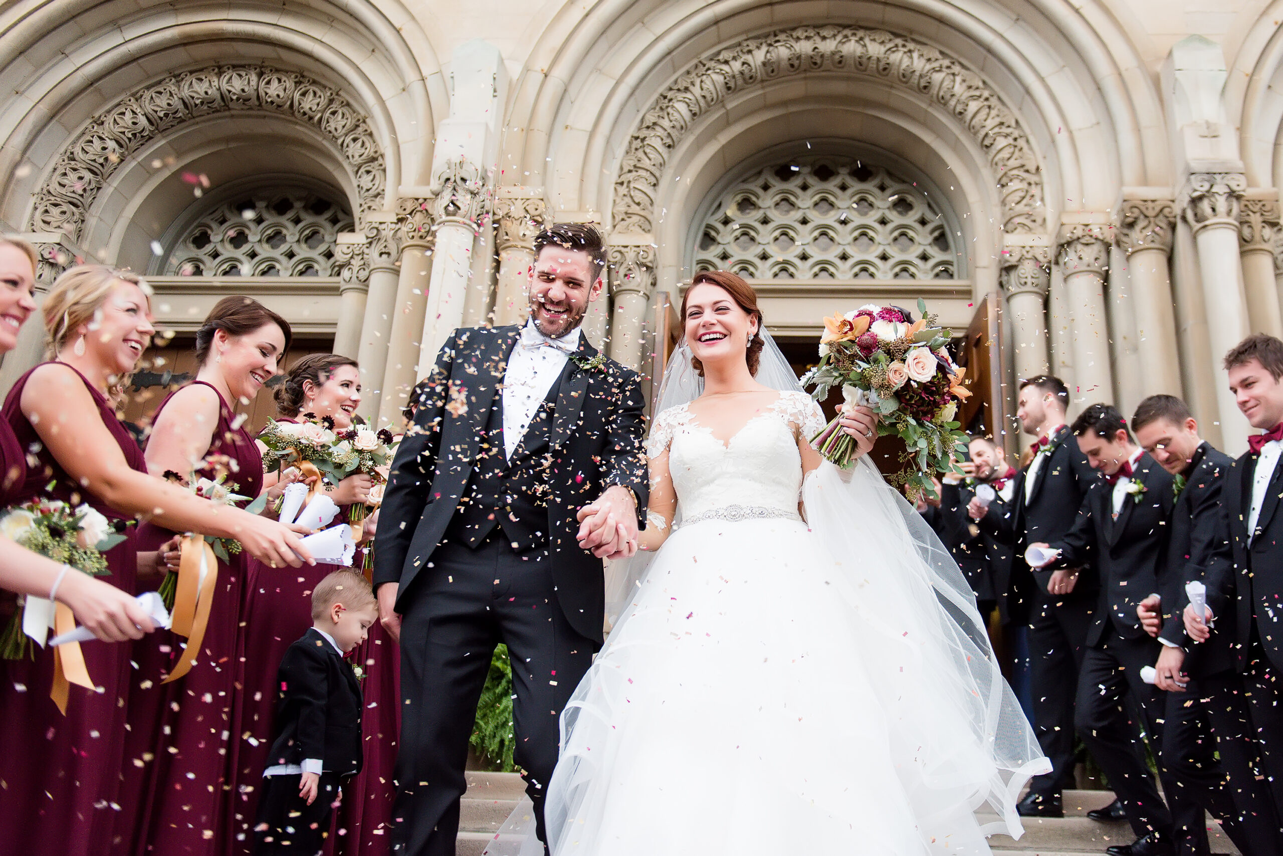 A photo of a bride and groom smiling and holding hands. Their wedding party threw rose petals into the air.
