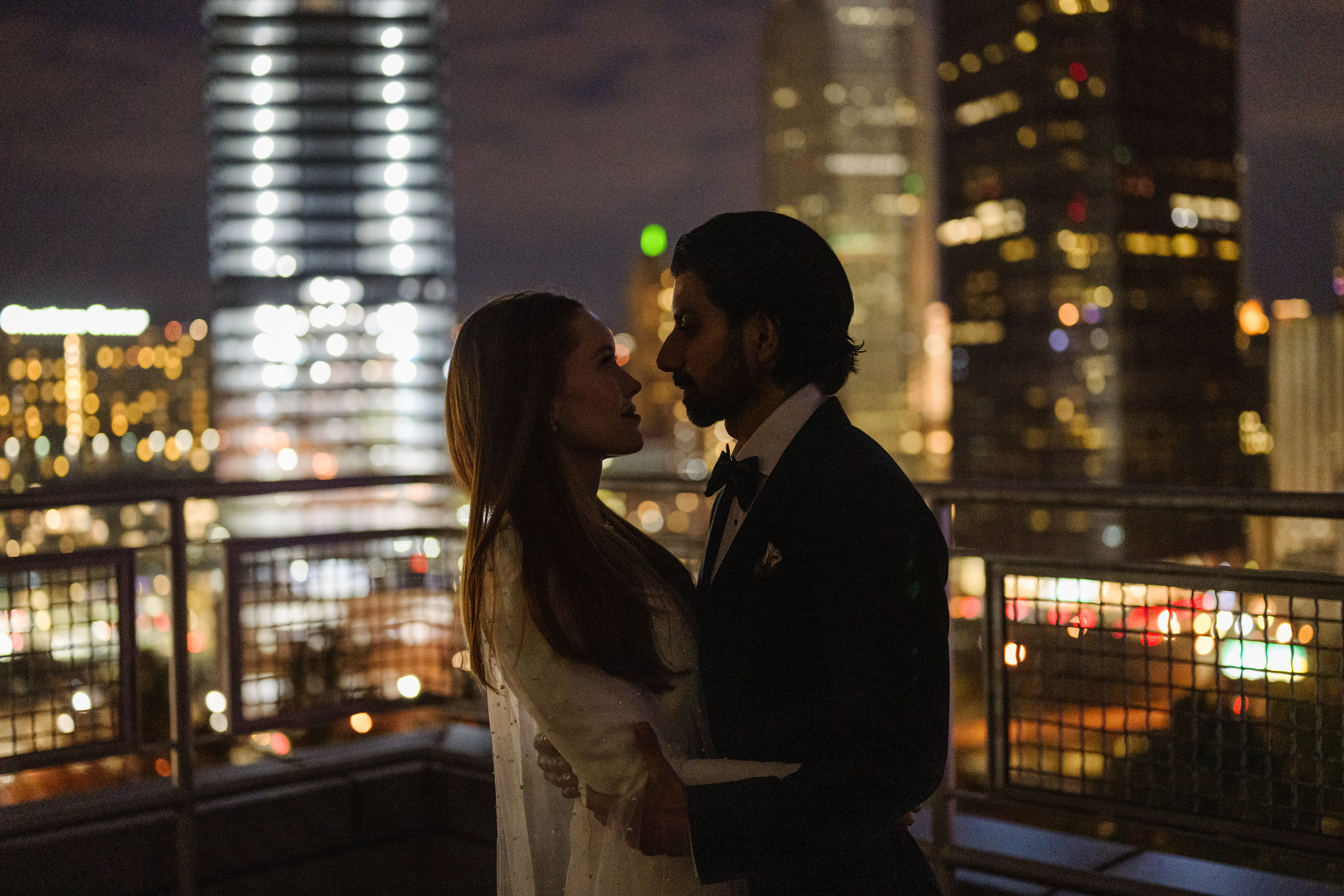 A darker photo of a bride and groom holding each other against a blurred backdrop of Pittsburgh downtown city lights.