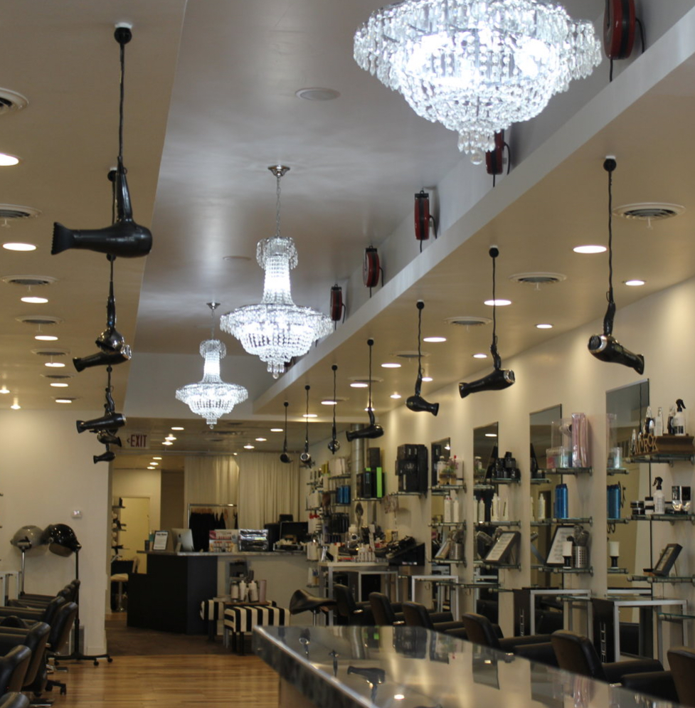 A photo of inside Sergio's Style. Blow dryers hang from the ceiling above each styling station, as well as chandeliers.
