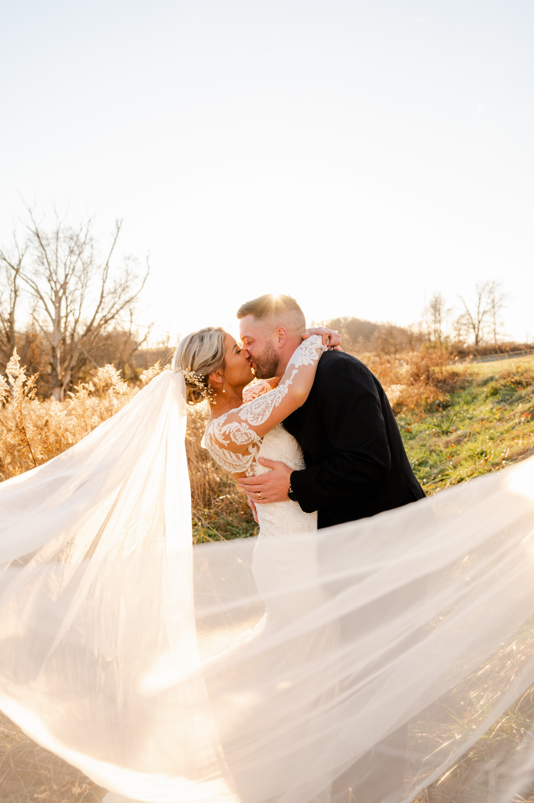 A photo of a bride and groom kissing outside with the bride's veil flowing in the wind. The sun shines bright behind the couple.