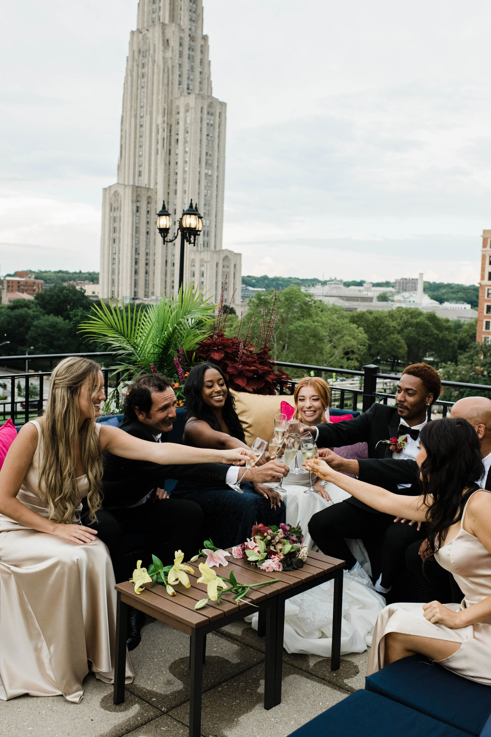 A photo of a wedding party clinking champagne glasses at the Rooftop Terrace at The University Club. The Cathedral of Learning and trees are in the background.