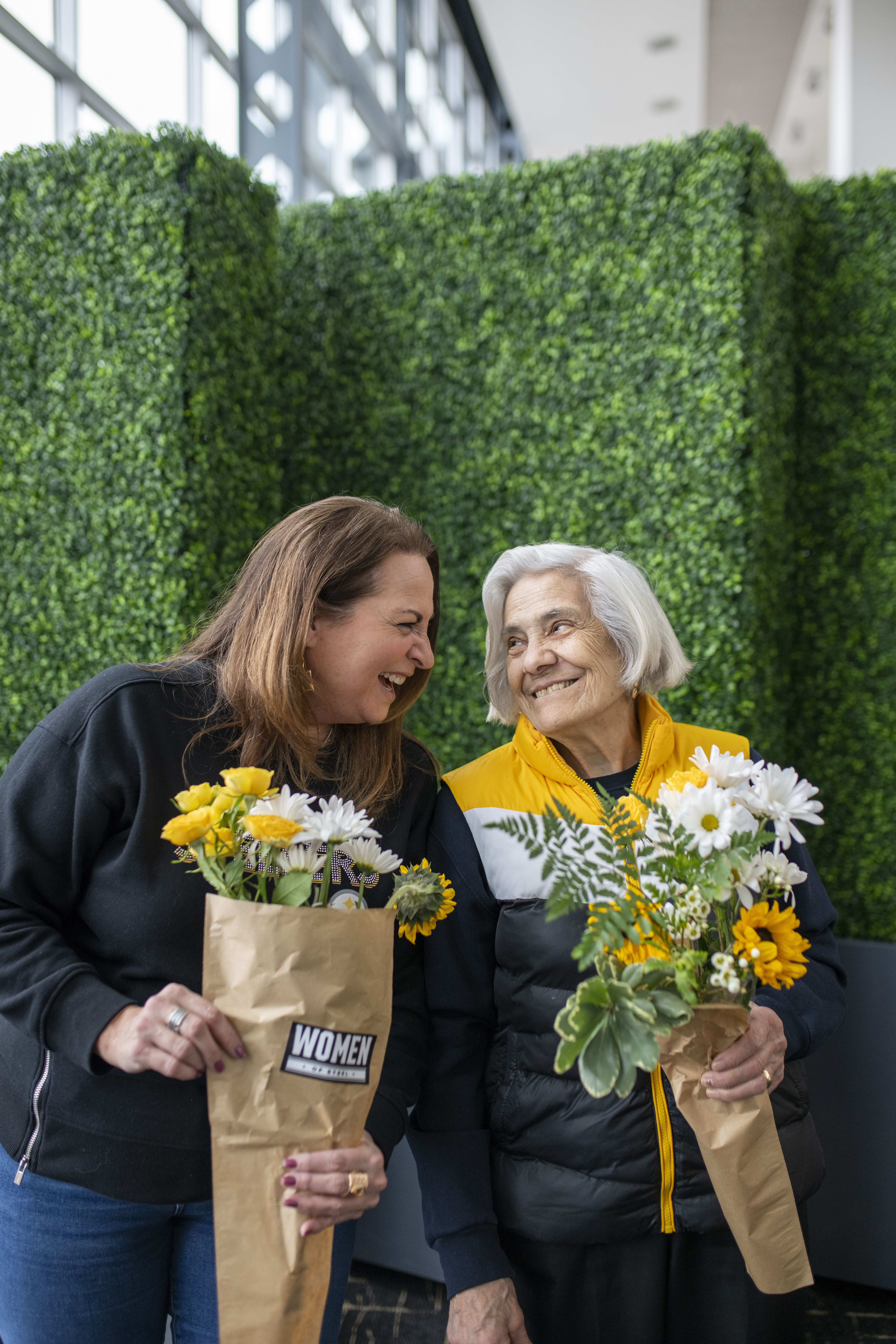 A photo of a middle aged woman and an elderly woman smiling at each other and holding white and yellow flowers at the SteelHERS Social event.