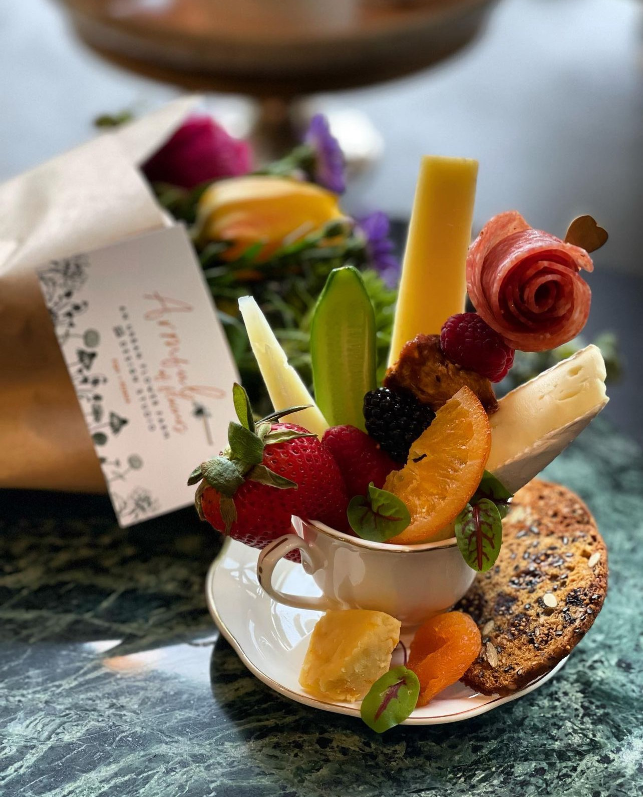 A photo of a food arrangement made by the Cheese Queen. A bouquet of fruits, cheeses, and meats sits in a teacup.