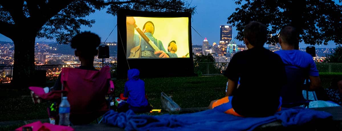 A photo of people sitting in Schenley Park Flagstaff Hill watching a movie outdoors, with the Pittsburgh skyline in the background.