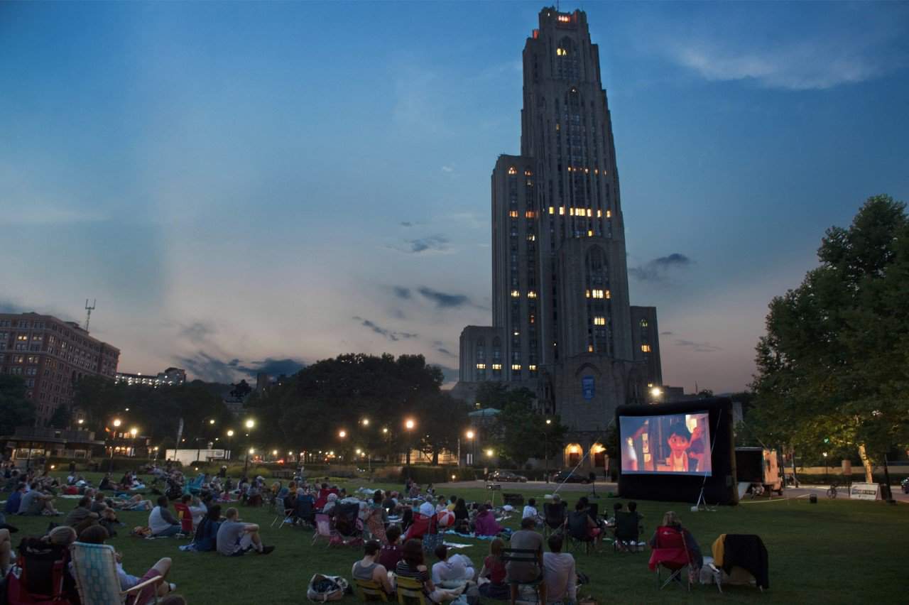 A photo of people sitting and watching a movie on the Schenley Plaza lawn at night, with the Cathedral of learning in the background.