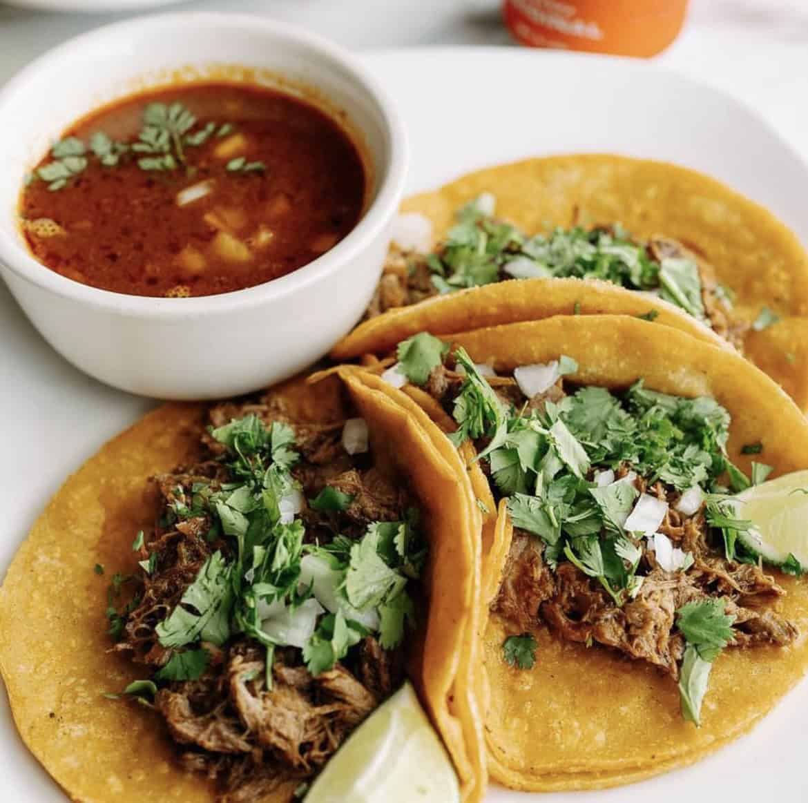 A photo of three tacos topped with onion and cilantro with a side of salsa.