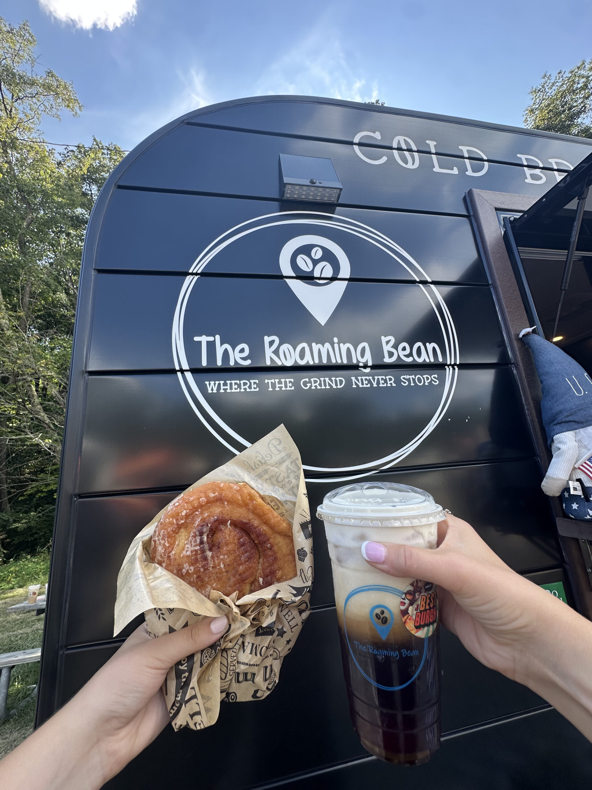 A photo of people holding a drink and bakery treat in front of the Roaming Bean sign.