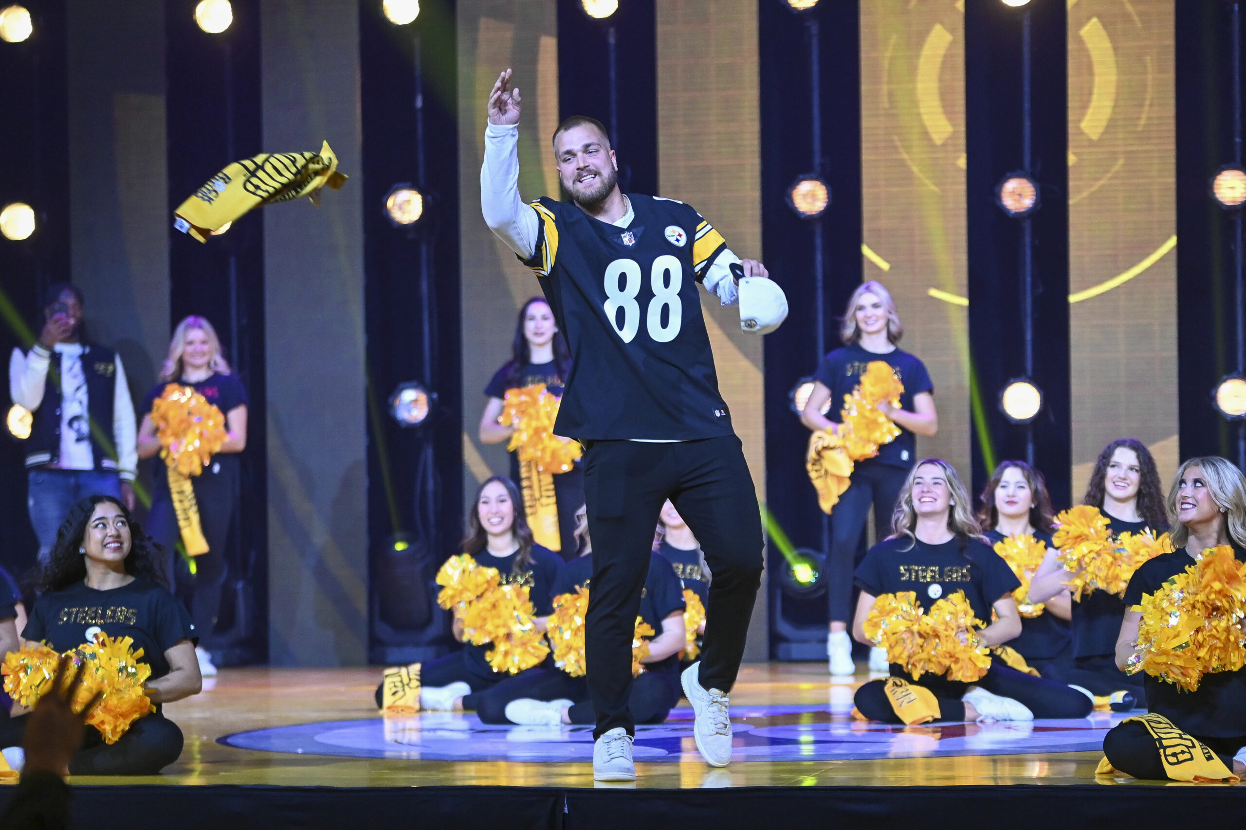 A photo of Pat Freiermuth, #88, in his jersey, throwing out Terrible Towels from a stage. Steelers cheerleaders sit and stand on the stage nearby.